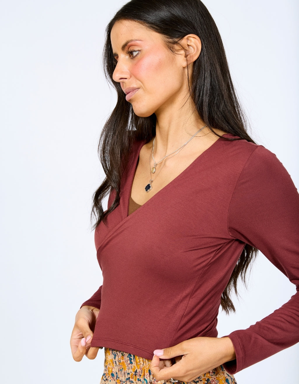 A woman wearing a long-sleeve wrap-style top in a muted burgundy or maroon color, paired with a patterned skirt. She is photographed against a clean, light gray studio background.