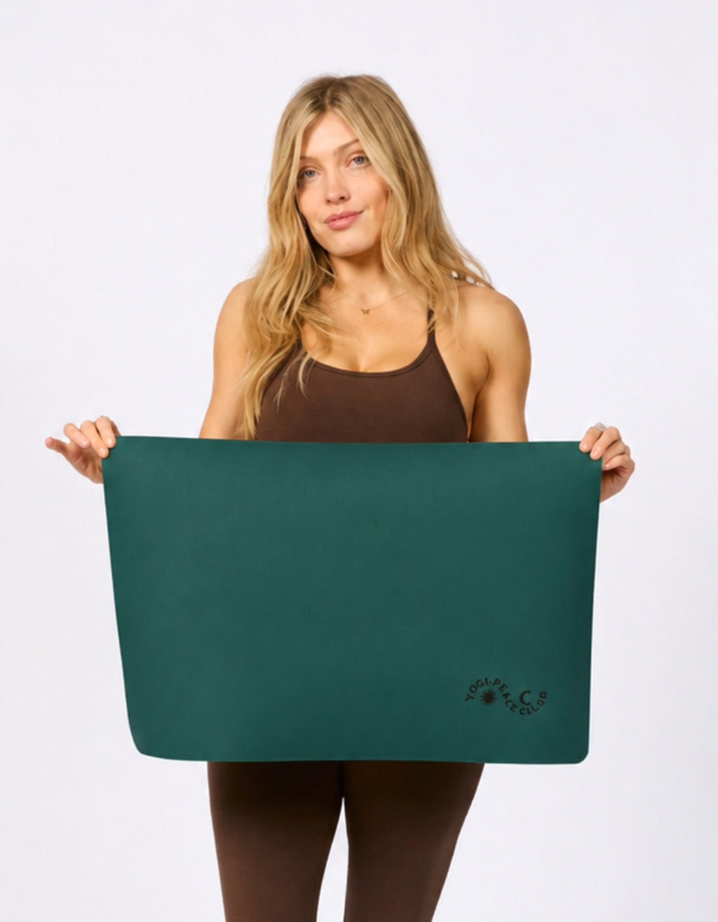 A woman holding a dark teal yoga mat against a plain white studio background. The yoga mat features a small etched logo in the bottom right corner .