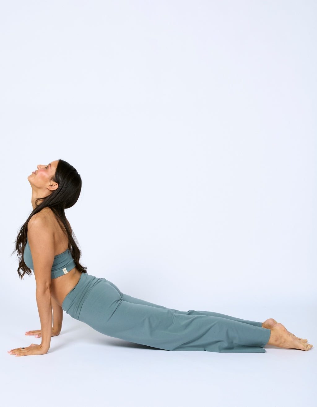 A woman in a matching sage green activewear set, consisting of a sports bra and high-waisted leggings, is performing a yoga pose on a seamless light gray studio background.
